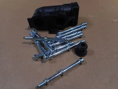 Schrauben und eine schwarze Kunststoffkomponente, möglicherweise von Remeha, liegen auf einer braunen Oberfläche. Die Schrauben sind metallisch glänzend und von mittlerer Länge.